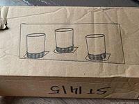 Cardboard box packaging shows an illustration of the candle holder with three candle cups mounted on a rectangular plaque.