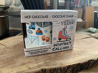 Front view of box showing part of the ceramic mug with winter-themed graphics, and text indicating maple hot chocolate mix included.