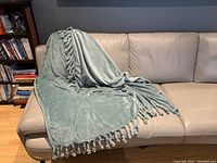 Light sage green oversized plush throw blanket with tassels draped over a beige leather couch.
