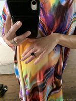 Photo showing the ring worn on a hand, featuring a large round black stone with a metallic band, against a colorful rainbow pattern shirt.