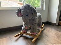 Front-side view of gray plush elephant rocking toy with wooden base on hardwood floor near window.