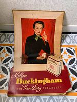 Frontal photo of vintage framed Buckingham cigarette advertisement showing woman holding cigarette and pack, ad is bent and damaged.
