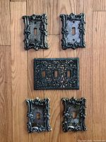 Top-down photo showing five vintage metal light switch covers on wooden floor: four single switch covers with ornate designs and one triple switch cover with filigree pattern.