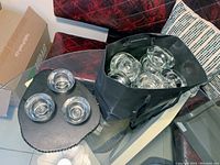 Three clear glass votive holders placed on a dark surface next to a bag filled with additional votive holders.