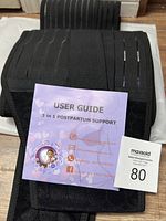 Photograph showing the black elastic postpartum support belt with ribbed texture and adjustable straps. A lavender-colored user guide titled '3 in 1 POSTPARTUM SUPPORT' and a small MaxSold auction card with lot number 80 are also visible.