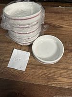 Six stacked white bone china bowls with a burgundy rim, wrapped in plastic, and one bowl placed aside unwrapped on a wooden surface