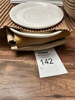 Stack of six white saucers/plates stacked on cardboard with a lot tag visible showing quantity