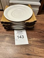 Stack of six white saucers/plates with circular pattern, on corrugated cardboard padding, showing lot number tag.