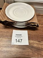 Stack of six white saucers on corrugated cardboard with auction label in front.