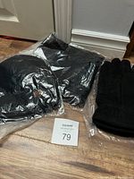 Top-down image of the three items in plastic packaging: toque, scarf, and gloves on wooden floor by white baseboard.