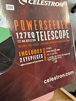 Box showing Celestron branding, PowerSeeker 127EQ model, 127 mm reflector telescope, includes 2 eyepieces and aluminum tripod with accessories.