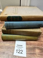 Four antique hardback books stacked horizontally, showing different colored covers and visible wear.