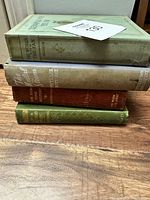 Four antique books stacked on a wooden surface, spines visible, titles partially readable.