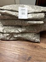 Stack of four folded light beige curtains with floral damask pattern, laid on wooden floor.