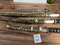 Three decorative swords arranged horizontally with ornate black and gold sheaths featuring dragon and floral embroidery, placed on a wooden floor.