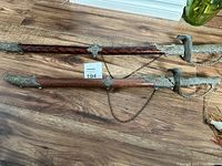 Two decorative swords shown sheathed, positioned side by side with visible ornate metal details on hilts and scabbards, braided tassels hanging from hilts.