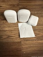 Three white wireless earbud charging cases on a wooden surface with a piece of paper marked '159'