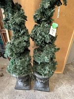 Front view of two artificial spiral trees with green foliage and string lights, in metal urn bases, standing side by side.