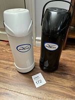 White and black Invicta insulated flasks standing side by side on the floor, showing full front view with logos visible.