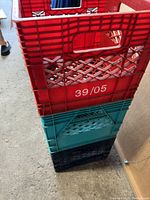 Stacked view of four plastic milk crates in red, teal, and black colors, showing wear and markings.