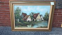 Frontal view of oil painting on canvas showing cottages by a lake in a gold decorative frame.