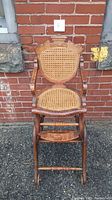 Front view of antique child's wooden high chair with cane seat and backrest, standing against a brick wall.
