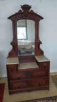 Front view of antique wooden dresser featuring ornate carved mirror frame, large central mirror, and marble top surfaces on drawers.