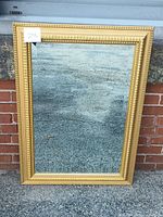 Front view of the rectangular gold gilt wall hanging mirror leaning against a brick wall outside.