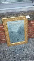 Full view of rectangular mirror with gold gilt ornate frame leaned against brick wall outdoors