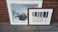 Two framed lithographs leaning against a brick wall; left is the Spencer William bald eagle, right is the Christina Miller birch tree scene