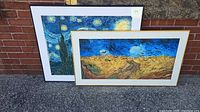 Two framed prints placed outdoors against a brick wall on pavement. The print with the Starry Night is in a black frame and positioned behind the Wheatfield with Cypresses print, which has a thinner light-colored frame.