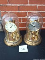 Two vintage anniversary clocks with glass domes and brass bases shown outdoors in front of a brick wall.