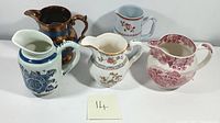 Front view of five ceramic pitchers showing various patterns and colors including blue floral, copper glazed, and red floral designs