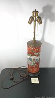Full view of the vintage porcelain lamp showing the brass light fixtures, porcelain body with hand-painted red and floral design, electrical cord, and wooden base.