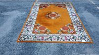 Full view of the rug laid flat on pavement showing overall color and design, predominantly rust-orange with cream border and medallions in center and corners.