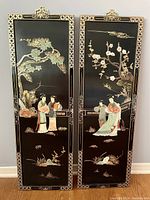 Pair of Asian Chinoiserie black lacquered wall panels featuring women and tree scenes accented with jade and mother of pearl inlay, viewed standing side by side on the floor against a light gray wall.