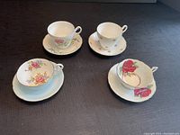 Four bone china teacups and saucers arranged on dark fabric surface, showing floral patterns and cup shapes