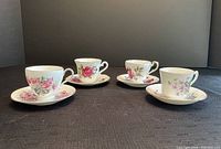 Four bone china teacups and matching saucers displayed on a dark surface. Each cup and saucer has distinct floral patterns.