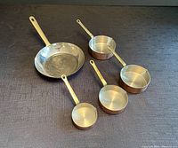 Five copper frying pan and pots arranged flat showing size and shape variety, brass handles visible, tinned interiors shiny.