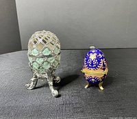 Front view of both decorative eggs showing the metal filigree glass egg on the left with music box interior and the blue and gold enamel egg on the right.