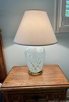 Glass table lamp with a light blue base decorated with raised white flowers and gold accents, topped with a white tapered fabric lampshade on a wooden surface.