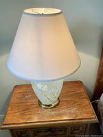 Full view of lamp with white tapered drum shade and floral glass base on wooden table.
