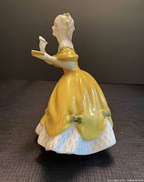 Side profile of the Royal Doulton figurine in yellow dress holding glass and tray.