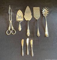 All seven silver-tone serving utensils laid out showing various sizes and styles including servers and cheese cutters.