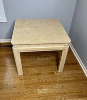 Square wood occasional table in a corner on a hardwood floor, showing front and side views. Table has light wood veneer top with patterned grain, visible wear spots and discoloration.