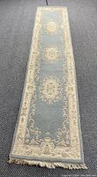 Full length view of carpet runner showing the blue field with three central cream colored medallions and ornate cream border with floral and scroll motifs, lying on a speckled dark carpet floor.