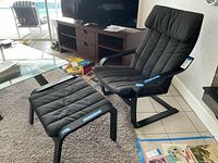 Black arm chair with matching ottoman shown from the side and front angles in a living room setting. Cushions show vertical stitching and appear lightly used.