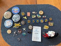 Top-down view of assortment of aviation and travel-related pins arranged on a dark woven placemat. Includes several Oshkosh year pins, airplane silhouettes, round and rectangular badges, and a small model airship.