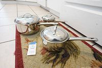 Set of three stainless steel pots and pans with lids shown on floor rug near white cabinetry. Pots have long handles and are shiny and reflective.