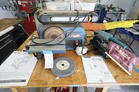 Ryobi belt/disc sander and Makita belt sander shown on a wooden table with manuals and a grinding disc.